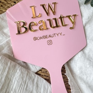 May include: A light pink handheld sign with a hexagonal top and a handle. The sign features the gold-colored raised text LW Beauty, the social media handle @LWBEAUTYY_, and an Instagram camera icon centered on the surface.