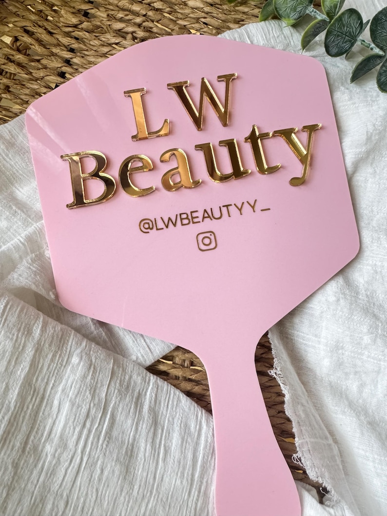 May include: A light pink handheld sign with a hexagonal top and a handle. The sign features the gold-colored raised text LW Beauty, the social media handle @LWBEAUTYY_, and an Instagram camera icon centered on the surface.