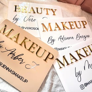K&ouml;nnte beinhalten: Vier rechteckige Acrylschilder mit erhabener goldfarbener Beschriftung. Die Schilder zeigen individuellen Text wie "BEAUTY by Vero", "MAKEUP By Adriana Barajas", "MAKEUP by Amber J" und "MAKEUP by Manar" sowie die dazugeh&ouml;rigen Social-Media-Namen.
