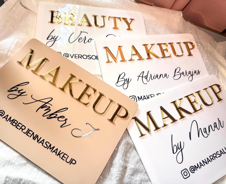 K&ouml;nnte beinhalten: Vier rechteckige Acrylschilder mit erhabener goldfarbener Beschriftung. Die Schilder zeigen individuellen Text wie "BEAUTY by Vero", "MAKEUP By Adriana Barajas", "MAKEUP by Amber J" und "MAKEUP by Manar" sowie die dazugeh&ouml;rigen Social-Media-Namen.