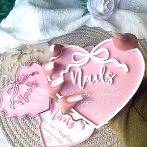 May include: Three pink heart-shaped acrylic signs with the word "Nails" in white lettering. The signs have white bows and a white border. The top sign says "Nails by Annabelle SHERMANDCO" and the bottom sign says "Nails by Zoe Chaplin CHAPLINNALSCO".