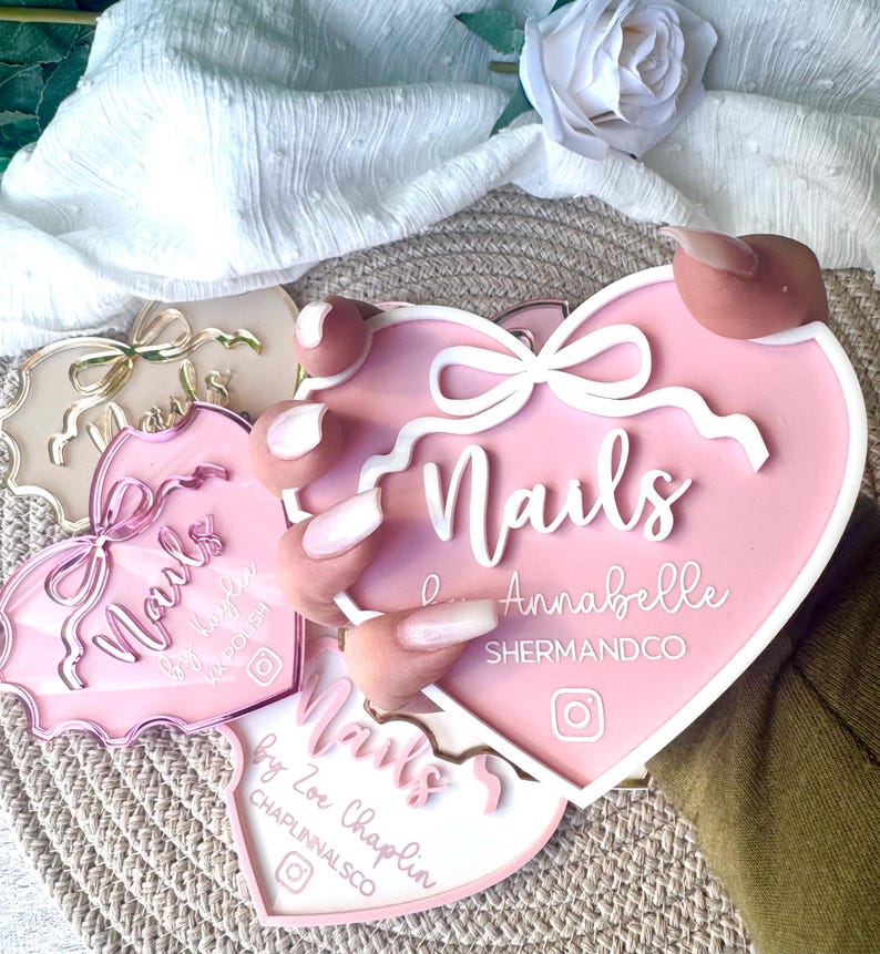 May include: Three pink heart-shaped acrylic signs with the word "Nails" in white lettering. The signs have white bows and a white border. The top sign says "Nails by Annabelle SHERMANDCO" and the bottom sign says "Nails by Zoe Chaplin CHAPLINNALSCO".