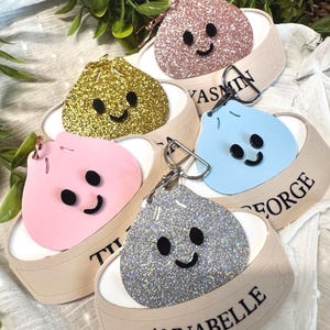 May include: Five keyrings in pink, gold, silver, blue, and pink, each with a smiling face and attached to a beige tag with a name printed on it. The keyrings are on a white surface with green leaves in the background.