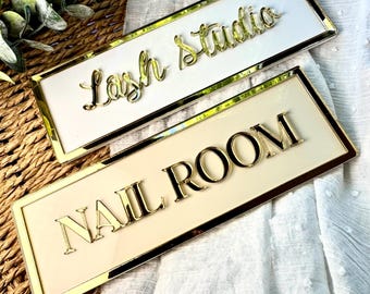 Puede incluir: Dos letreros rectangulares con bordes y letras doradas. El letrero superior dice "Lash Studio" en letras doradas cursivas sobre un fondo blanco. El letrero inferior dice "NAIL ROOM" en letras doradas en bloque sobre un fondo crema.