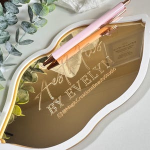 May include: Gold mirror in the shape of a lip with the text "Aesthetics by Evelyn" and the Instagram handle "@MagicCreationsBeautyStudio". A pink and white pen is resting on the mirror.