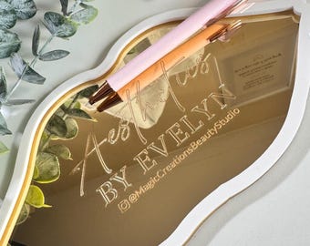 May include: Gold mirror in the shape of a lip with the text "Aesthetics by Evelyn" and the Instagram handle "@MagicCreationsBeautyStudio". A pink and white pen is resting on the mirror.