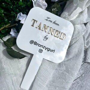 May include: White hand mirror with a gold inscription that reads "I've been tanned by @Bronzygurl".