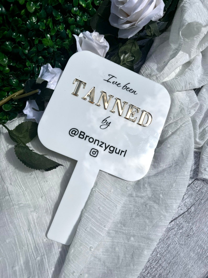 May include: White hand mirror with a gold inscription that reads "I've been tanned by @Bronzygurl".