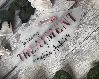 May include: A white marble-patterned sign with the text "I've had my TREATMENT at Rosabela Aesthetics" in black and rose gold lettering. A small rose gold syringe is placed on the sign.