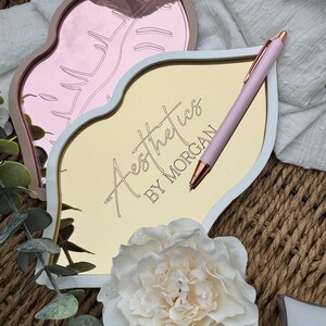 May include: A white and gold lip-shaped tray with the text "Aesthetics by Morgan" on the front. A pink pen is lying on the tray. The tray is sitting on a woven surface.
