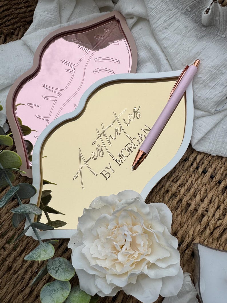 May include: A white and gold lip-shaped tray with the text "Aesthetics by Morgan" on the front. A pink pen is lying on the tray. The tray is sitting on a woven surface.