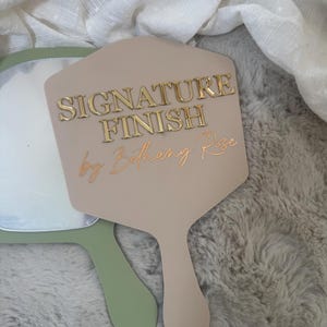 May include: A beige handheld mirror with a hexagonal shape featuring the gold embossed text SIGNATURE FINISH and the script text by Bethany Rose. A second sage green handheld mirror is partially visible underneath the beige mirror.