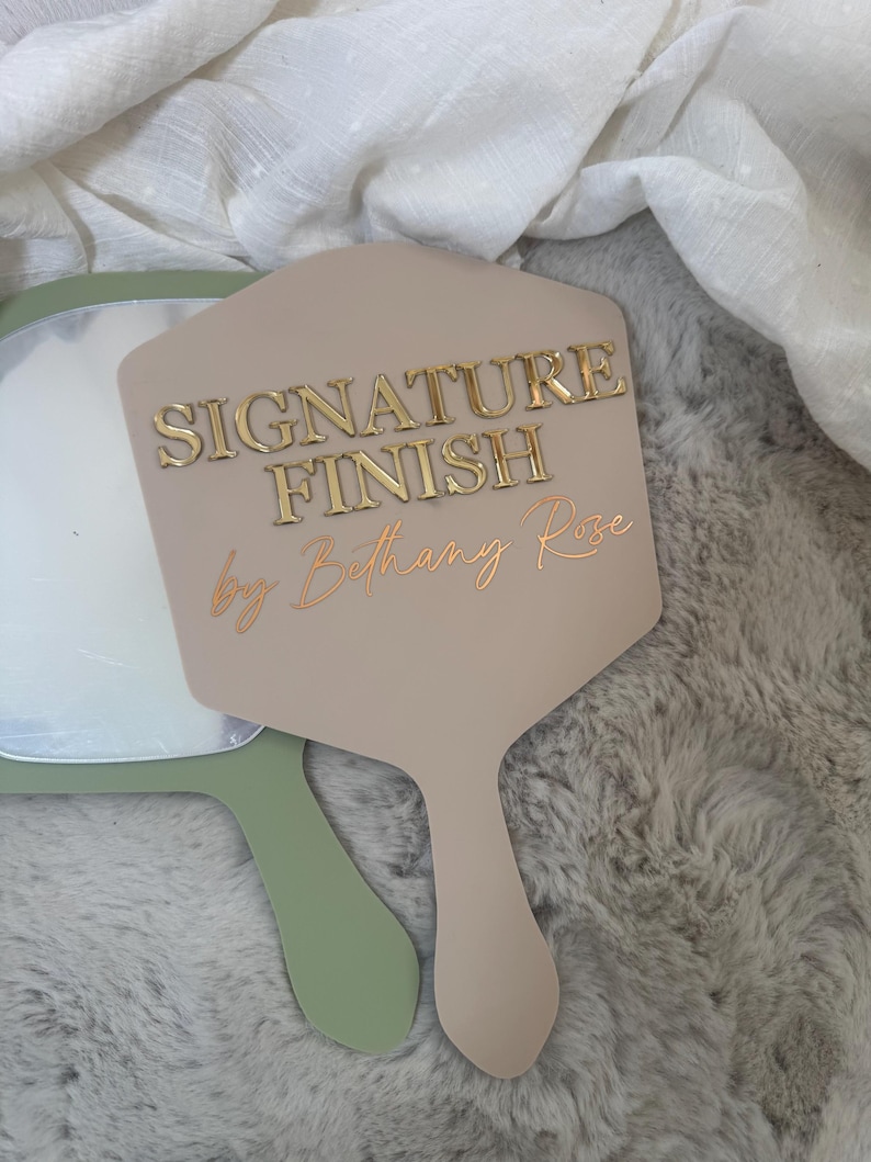 May include: A beige handheld mirror with a hexagonal shape featuring the gold embossed text SIGNATURE FINISH and the script text by Bethany Rose. A second sage green handheld mirror is partially visible underneath the beige mirror.