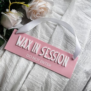 May include: A pink rectangular sign with the words "WAX IN SESSION" and "DO NOT ENTER" in white, with a white ribbon attached. The sign is set against a white textured background, with white roses and greenery in the background.
