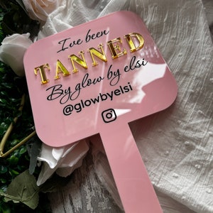 May include: Pink acrylic sign with the text "I've been TANNED By glow by elsi @glowbyelsi" in black and gold lettering. The sign has a handle and an Instagram icon.