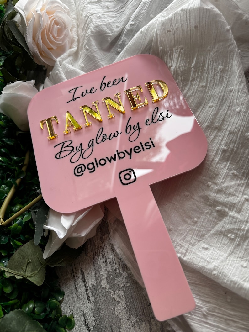 May include: Pink acrylic sign with the text "I've been TANNED By glow by elsi @glowbyelsi" in black and gold lettering. The sign has a handle and an Instagram icon.