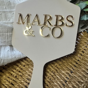 May include: A beige, paddle-shaped sign featuring the gold, reflective, three-dimensional text MARBS & CO centered on the surface. The sign has a smooth, matte finish and a handle at the bottom, presenting a clean, professional appearance.