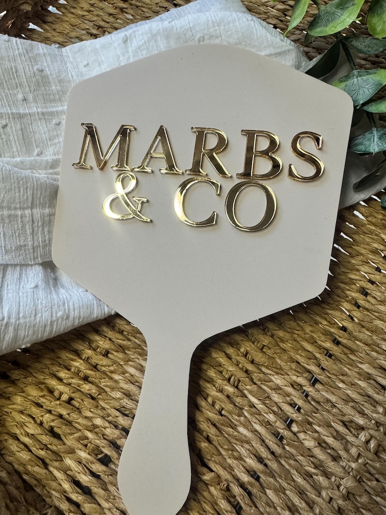 May include: A beige, paddle-shaped sign featuring the gold, reflective, three-dimensional text MARBS & CO centered on the surface. The sign has a smooth, matte finish and a handle at the bottom, presenting a clean, professional appearance.