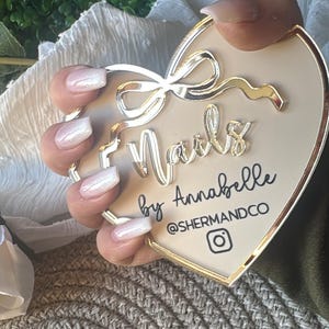 May include: A white heart-shaped sign with gold trim and a gold bow. The sign says "Nails by Annabelle @shermandco" with an Instagram icon. A hand with white ombre nails is holding the sign.