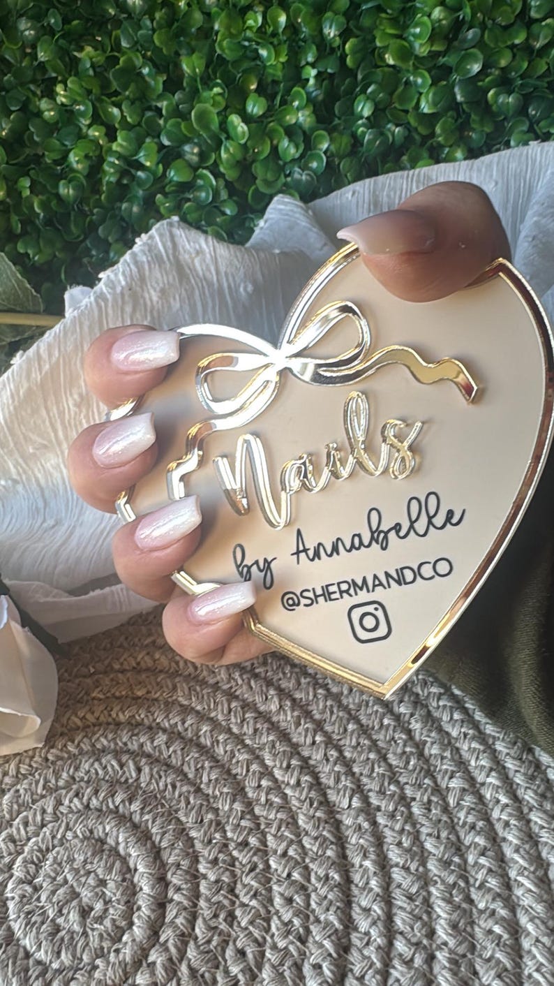 May include: A white heart-shaped sign with gold trim and a gold bow. The sign says "Nails by Annabelle @shermandco" with an Instagram icon. A hand with white ombre nails is holding the sign.