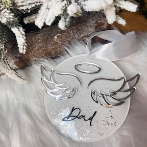 May include: White round ornament with silver angel wings and a halo. The ornament has a glittery background and the word "Dad" written in black.