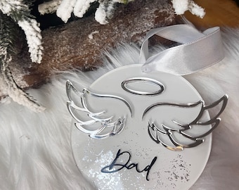 May include: White round ornament with silver angel wings and a halo. The ornament has a glittery background and the word "Dad" written in black.