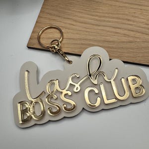 May include: A white acrylic keychain with a gold glitter finish. The keychain features the text "Lash Boss Club" in a gold glitter font.