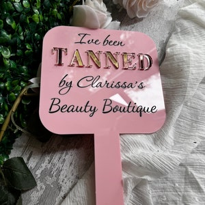 May include: Pink acrylic sign with the text "I've been TANNED by Clarissa's Beauty Boutique" in gold letters.
