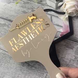 May include: A beige hand mirror with a gold-colored syringe design and the text "Flawless Aesthetics by Lucy".