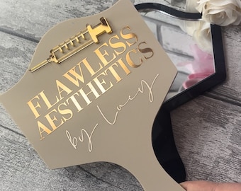 May include: A beige hand mirror with a gold-colored syringe design and the text "Flawless Aesthetics by Lucy".