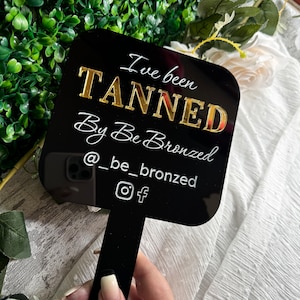 May include: Black square sign with gold lettering that reads "I've been TANNED By Be Bronzed @ _be_bronzed" with Instagram and Facebook icons.