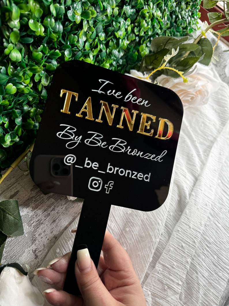 May include: Black square sign with gold lettering that reads "I've been TANNED By Be Bronzed @ _be_bronzed" with Instagram and Facebook icons.