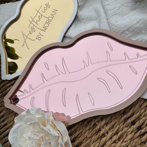 May include: Two pink and gold lip-shaped trays with mirrored surfaces. The gold tray has the text "Aesthetics by Morgan" engraved on it.
