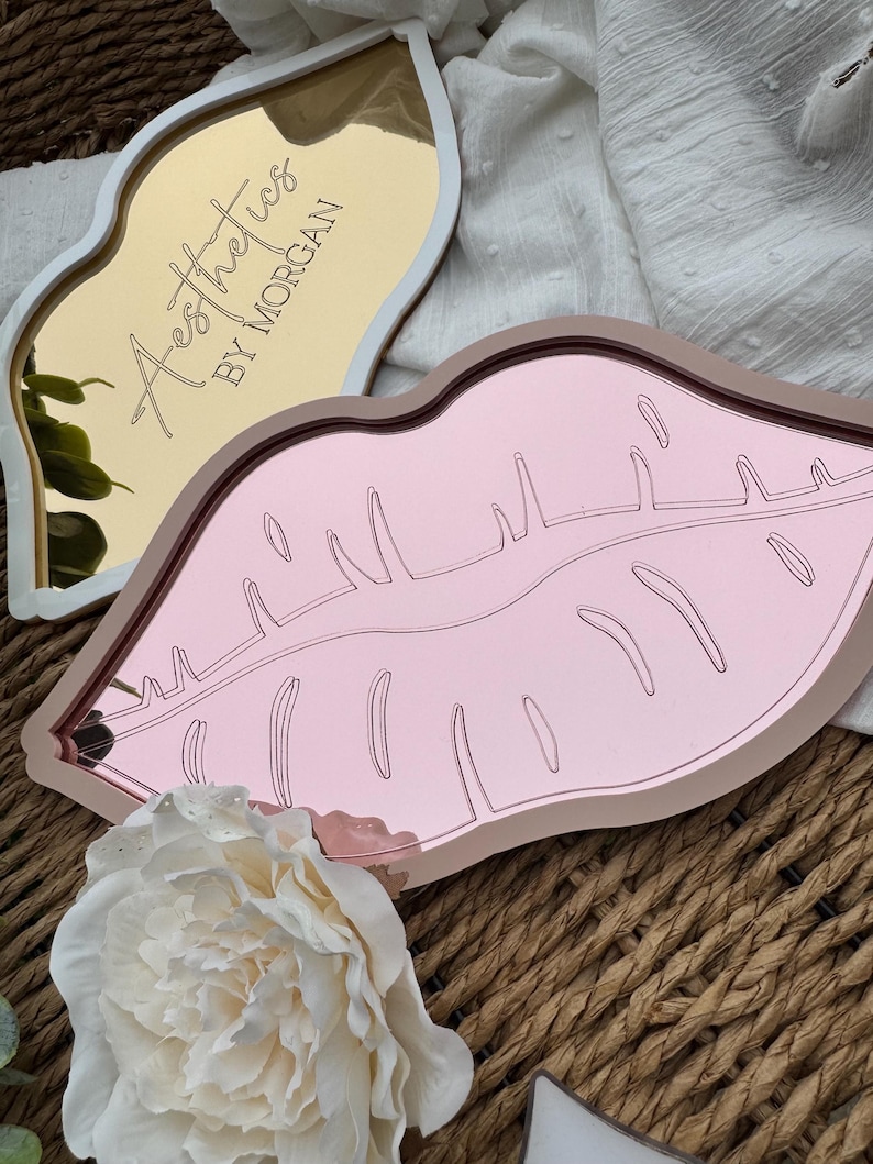 May include: Two pink and gold lip-shaped trays with mirrored surfaces. The gold tray has the text "Aesthetics by Morgan" engraved on it.