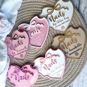 May include: Five pink, white and gold heart-shaped acrylic signs with the word "Nails" and the name of the nail artist. Each sign has a gold ribbon and a small Instagram icon.
