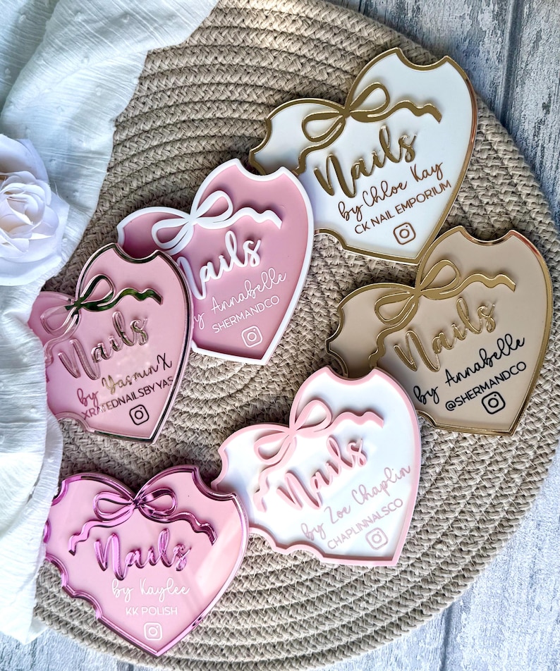 May include: Five pink, white and gold heart-shaped acrylic signs with the word "Nails" and the name of the nail artist. Each sign has a gold ribbon and a small Instagram icon.