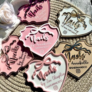 May include: Five pink heart-shaped acrylic nail art display stands with gold accents. Each stand features a white background with the text "Nails" and the name of the nail artist. The stands are arranged in a circle on a woven surface.