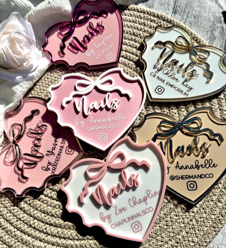 May include: Five pink heart-shaped acrylic nail art display stands with gold accents. Each stand features a white background with the text "Nails" and the name of the nail artist. The stands are arranged in a circle on a woven surface.