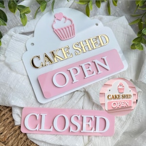 Op de afbeelding: Een wit acrylbord met een roze cupcake-icoon met een hartje, de gouden tekst CAKE SHED en een verwijderbaar roze plaatje met de witte tekst OPEN. Een apart roze rechthoekig plaatje met de witte tekst CLOSED is inbegrepen.