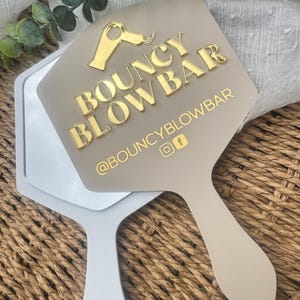 May include: A beige handheld mirror featuring a gold acrylic icon of a hair dryer and the text BOUNCY BLOWBAR in gold lettering. Below the text, the handle area displays the social media handle @BOUNCYBLOWBAR with Instagram and Facebook icons.