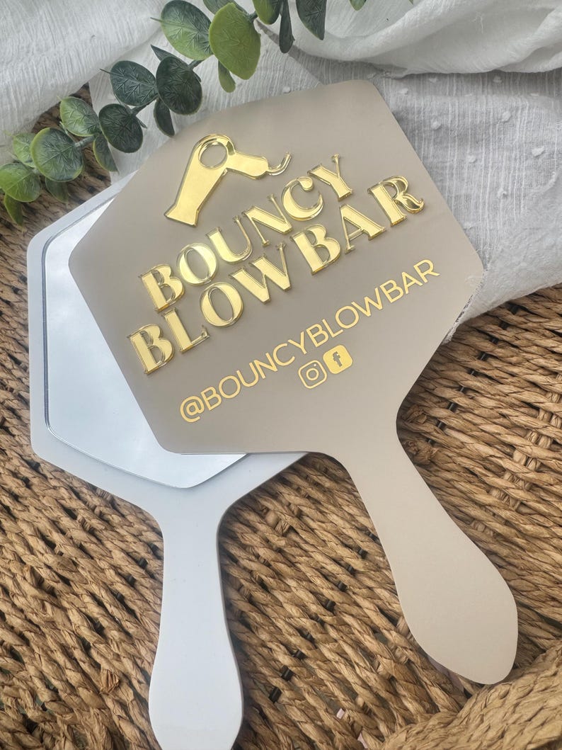 May include: A beige handheld mirror featuring a gold acrylic icon of a hair dryer and the text BOUNCY BLOWBAR in gold lettering. Below the text, the handle area displays the social media handle @BOUNCYBLOWBAR with Instagram and Facebook icons.