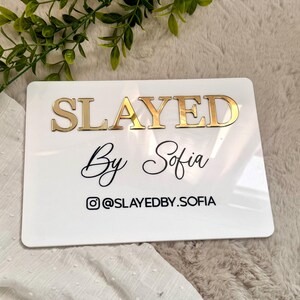 K&ouml;nnte beinhalten: Ein rechteckiges wei&szlig;es Acrylschild mit dem Wort SLAYED in erhabenen, goldfarbenen Metallbuchstaben. Darunter steht der Text By Sofia in schwarzer Schreibschrift, gefolgt von einem Instagram-Symbol und dem Benutzernamen @SLAYEDBY.SOFIA in Schwarz.