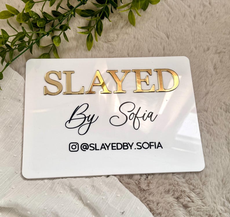 K&ouml;nnte beinhalten: Ein rechteckiges wei&szlig;es Acrylschild mit dem Wort SLAYED in erhabenen, goldfarbenen Metallbuchstaben. Darunter steht der Text By Sofia in schwarzer Schreibschrift, gefolgt von einem Instagram-Symbol und dem Benutzernamen @SLAYEDBY.SOFIA in Schwarz.