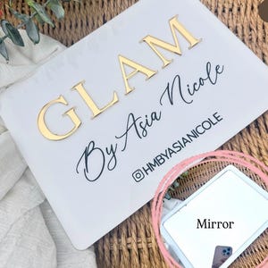K&ouml;nnte beinhalten: Ein rechteckiges wei&szlig;es Schild mit dem goldenen 3D-Schriftzug GLAM und dem schwarzen Schreibschrifttext By Asia Nicole, darunter der Instagram-Name @HMBYASIANICOLE. Ein kleiner rechteckiger Spiegel befindet sich in der unteren rechten Ecke.
