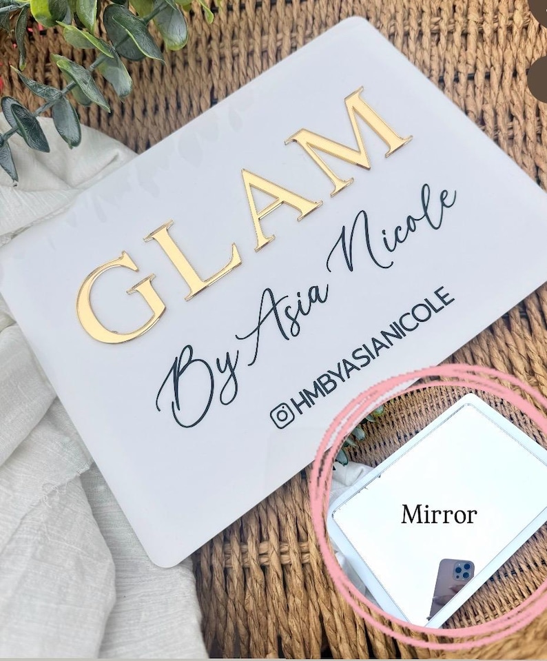 K&ouml;nnte beinhalten: Ein rechteckiges wei&szlig;es Schild mit dem goldenen 3D-Schriftzug GLAM und dem schwarzen Schreibschrifttext By Asia Nicole, darunter der Instagram-Name @HMBYASIANICOLE. Ein kleiner rechteckiger Spiegel befindet sich in der unteren rechten Ecke.
