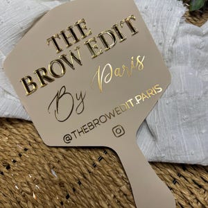May include: A beige hand mirror with gold lettering that reads "THE BROW EDIT By Paris". The mirror has a handle and an Instagram handle printed on it. The mirror is on a woven surface.