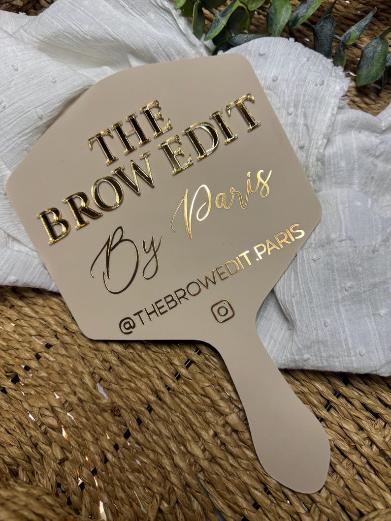 May include: A beige handheld mirror featuring the gold text THE BROW EDIT By Paris and the Instagram handle @THEBROWEDIT.PARIS with an Instagram icon. The mirror has a flat, paddle-shaped design with a smooth, matte finish surface.