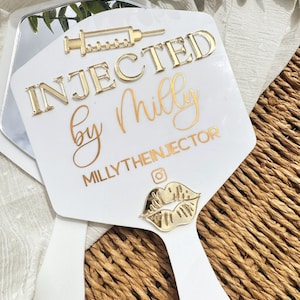 May include: A white hand mirror with gold text that reads "INJECTED by Milly" and "MILLYTHEINJECTOR". The mirror has a gold syringe and lip design. The mirror is on a woven surface.