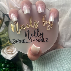 Puede incluir: Primer plano de una mano con uñas blancas con degradado, sosteniendo un posavasos redondo blanco con el texto "Nails by Nelly @NEL_LYNAILZ" en letras doradas.
