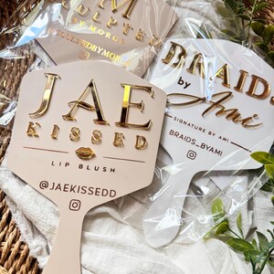 May include: Three handheld hexagonal mirrors with custom gold-toned lettering. One reads JAE KISSED LIP BLUSH @JAEKISSEDD, another BRAIDS BY AMI SIGNATURE BY AMI BRAIDS_BYAMI, and the third SCULPTED BY MORGS SCULPTEDBYMORGS, all encased in clear plastic.